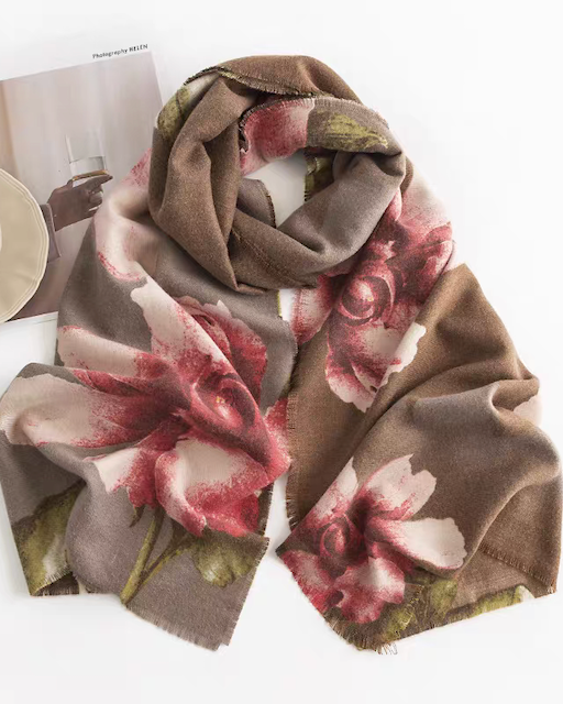 A long scarf with a large floral print in dusty pink, red, and muted green on a taupe-brown base, styled in a flat lay with a coffee cup and magazine on a white background.