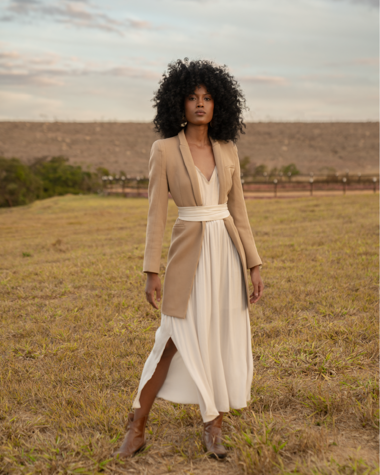 Woman in a beige coat and white dress standing in a field with a cloudy sky.