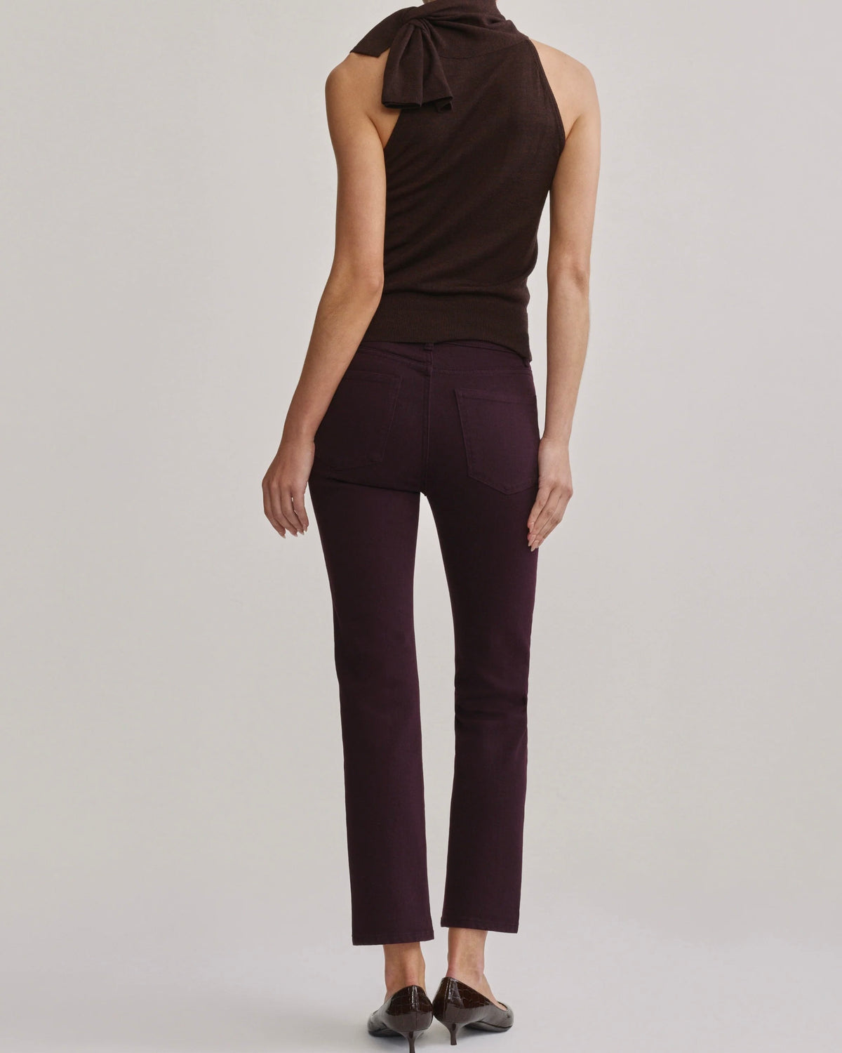 A model shown from the back wearing dark plum-colored ankle-length pants and a dark brown sleeveless top with a bow detail at the neck, set against a plain light gray studio background.