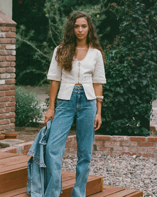 Woman in a white blouse and blue jeans standing outdoors with greenery in the background