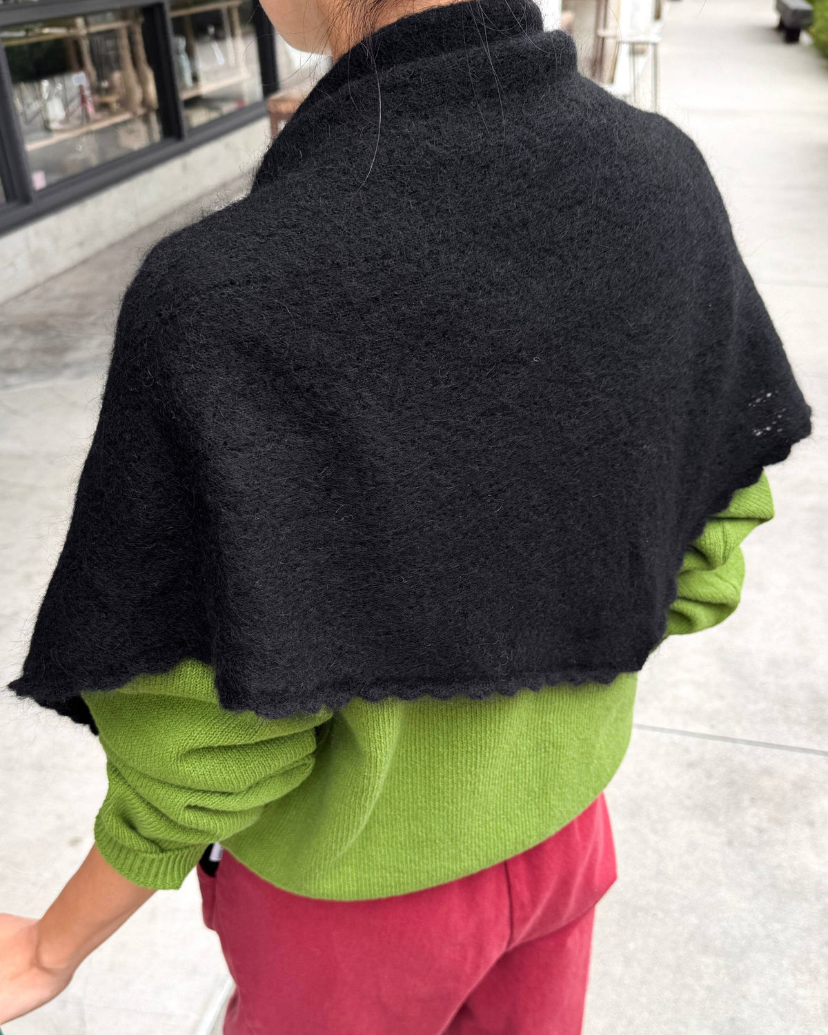 A person viewed from behind wearing a black fuzzy knit shawl or cape over a bright green sweater and pink pants, standing outdoors on a paved walkway.