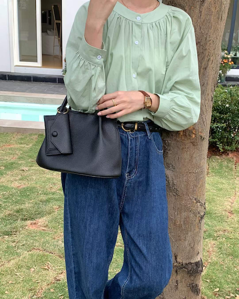 Person wearing a light green blouse and blue jeans, holding a black handbag outdoors.