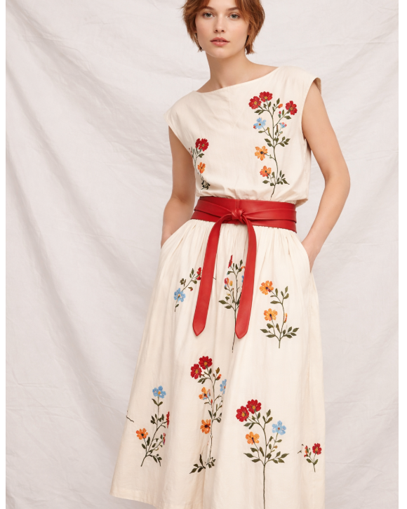 A woman modeling a sleeveless cream-colored dress with colorful floral embroidery and a wide red waist tie, set against a draped white fabric backdrop.