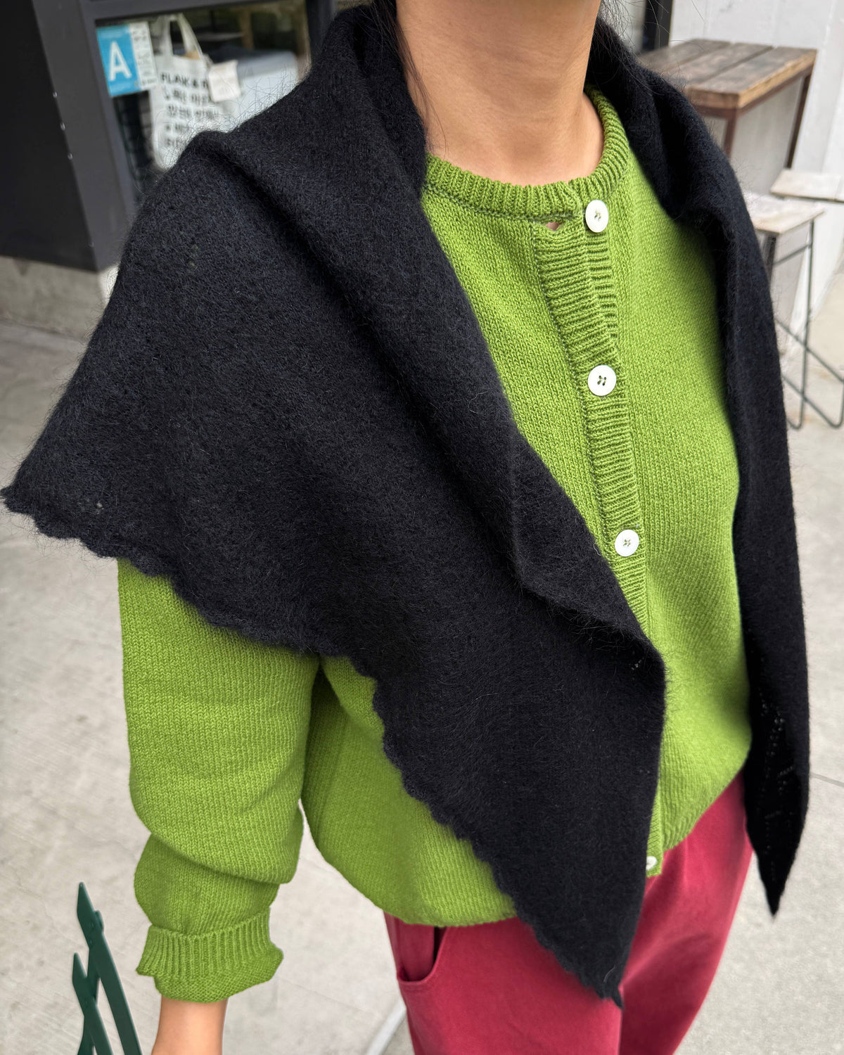 A person wearing a bright lime green knit cardigan with white buttons, layered with a black triangular shawl draped over the shoulders, paired with magenta pants.