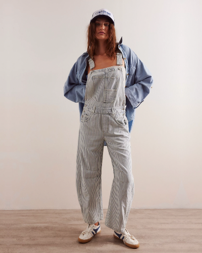 Model wearing the Free People Good Luck Stripe Overall in indigo railroad, standing on a wood floor in front of a white wall. Overalls are styled with denim button down shirt and a trucker hat.  