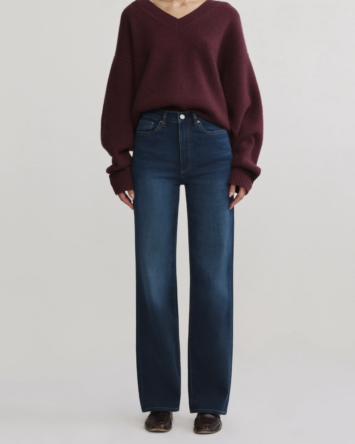 A model wearing a dark maroon knit V-neck sweater and dark blue wide-leg jeans, standing against a light gray studio background.