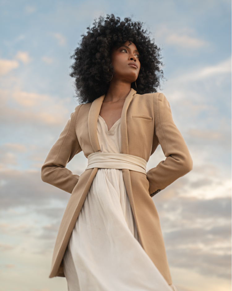 Woman wearing a beige coat over a white dress against a cloudy sky.