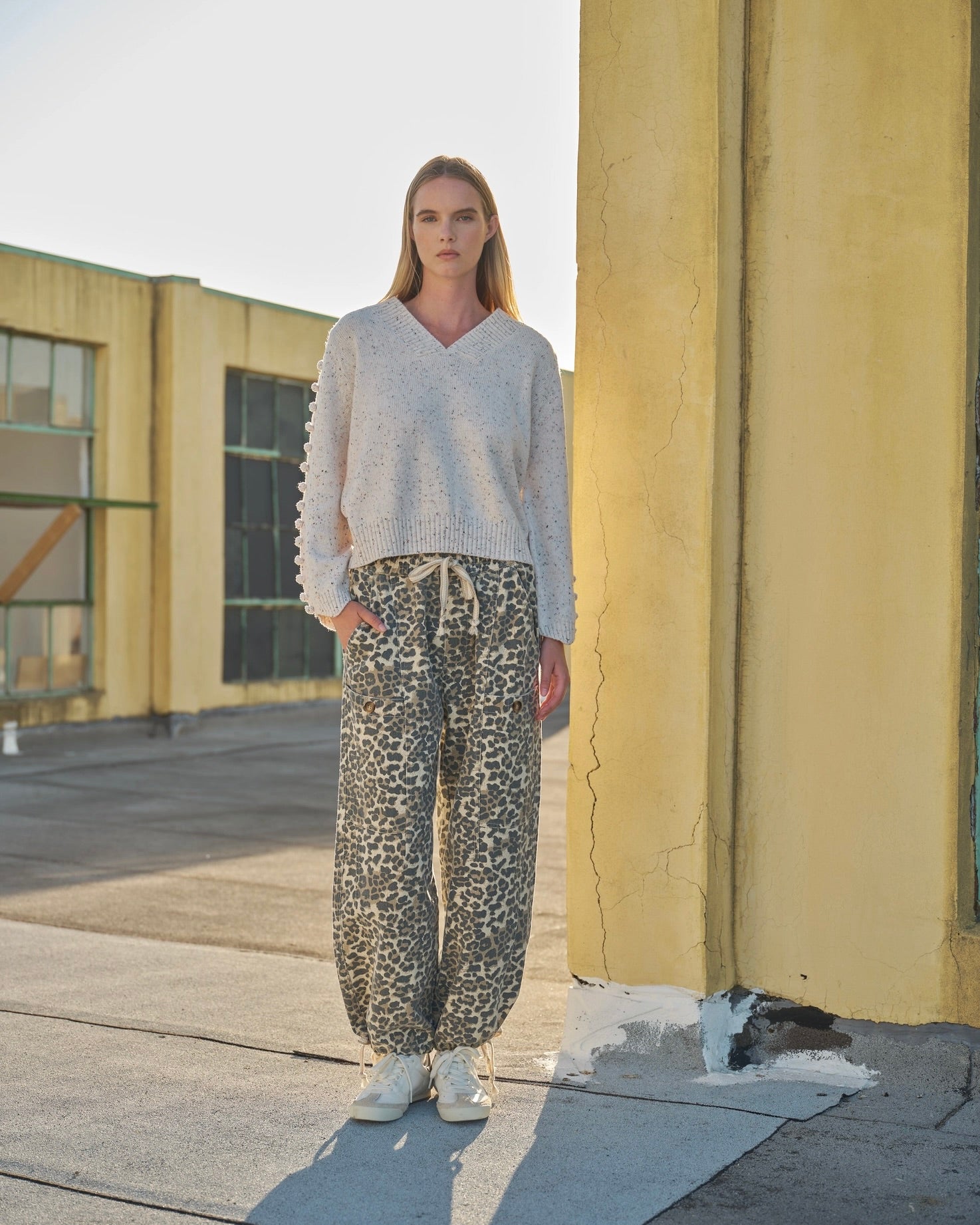 Model standing outside wearing the Aaron & Amber  relaxed leopard print pull on denim pant wearing a beige sweater with white sneakers.