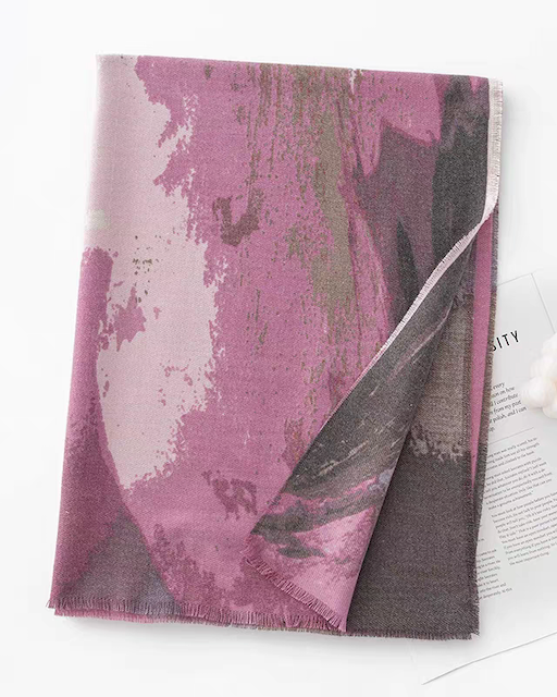 A folded rectangular scarf or throw with a painterly abstract pattern in dusty pink, mauve, and charcoal gray, styled on a white surface with a small decorative paper and white pom-pom accessory nearby.
