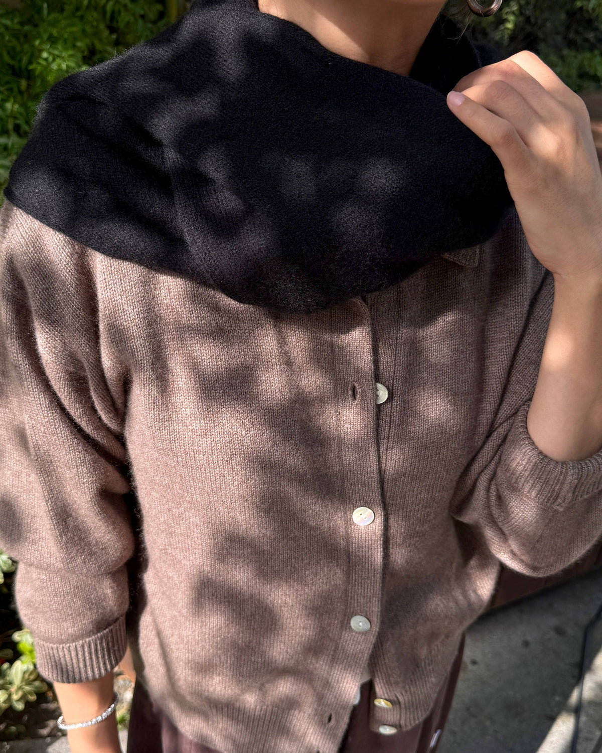 A person outdoors wearing a taupe knit button-front cardigan with a large black fuzzy collar or neck warmer draped around the shoulders.