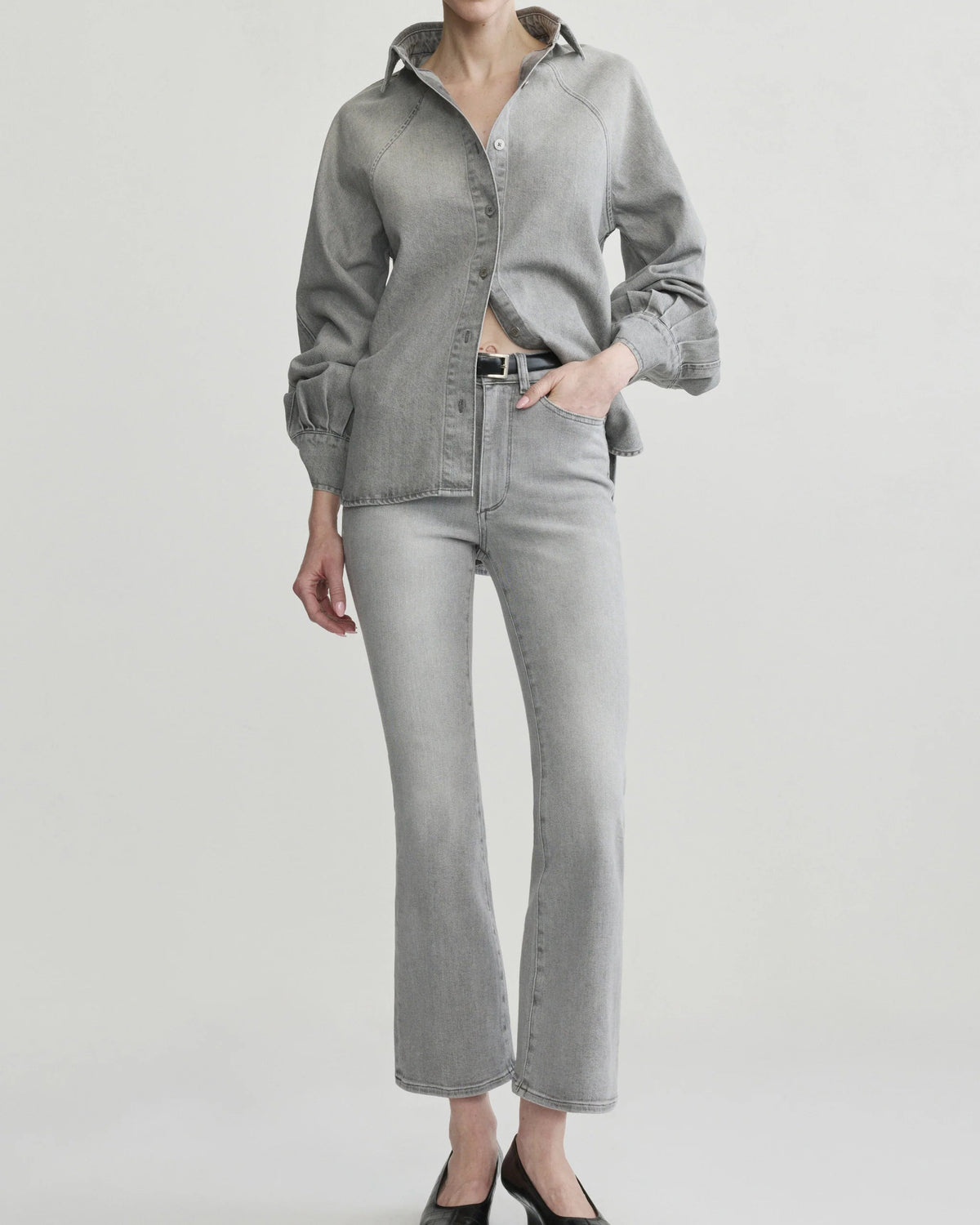 A model wearing a light gray denim button-up shirt and matching light gray denim flare jeans, paired with black pointed-toe heels, against a plain light gray studio background.