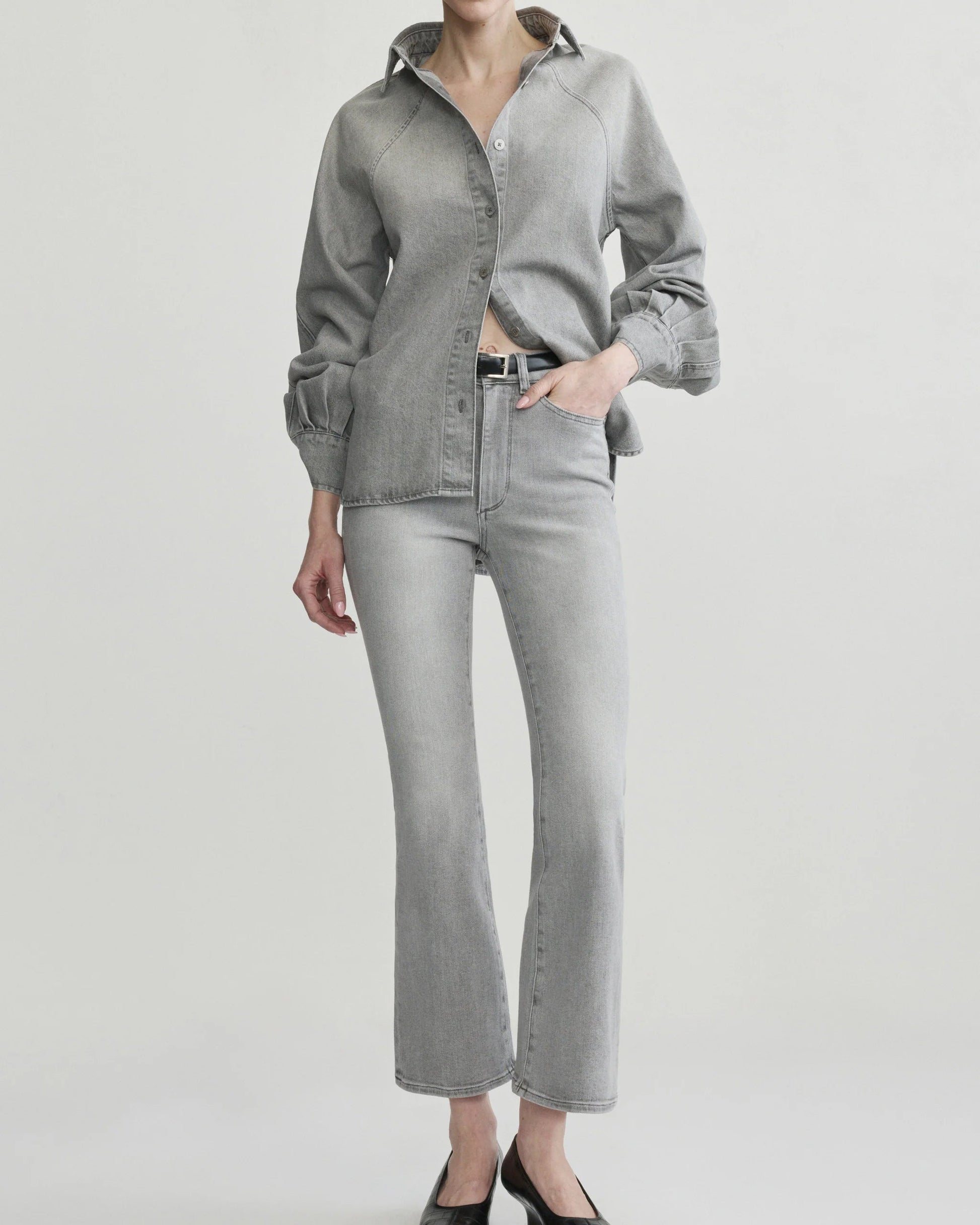 A model wearing a light gray denim button-up shirt and matching light gray denim flare jeans, paired with black pointed-toe heels, against a plain light gray studio background.