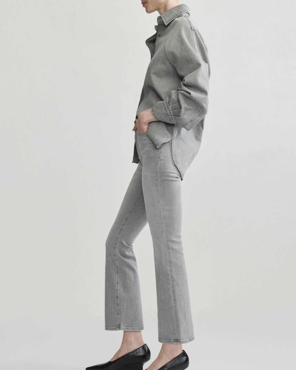 A model shown from the side wearing a light gray button-up shirt, matching light gray flared jeans, and black pointed-toe wedge pumps against a plain light gray studio background.