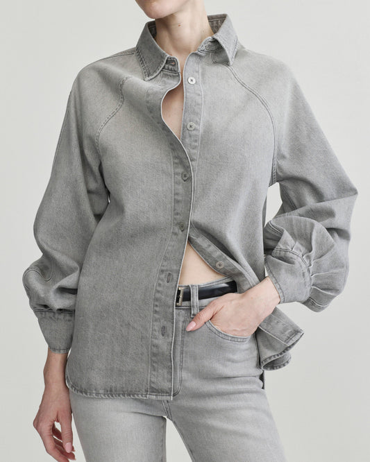 A model wearing a light gray washed denim button-up shirt with unique raglan sleeves and matching gray jeans, photographed against a solid light gray background.
