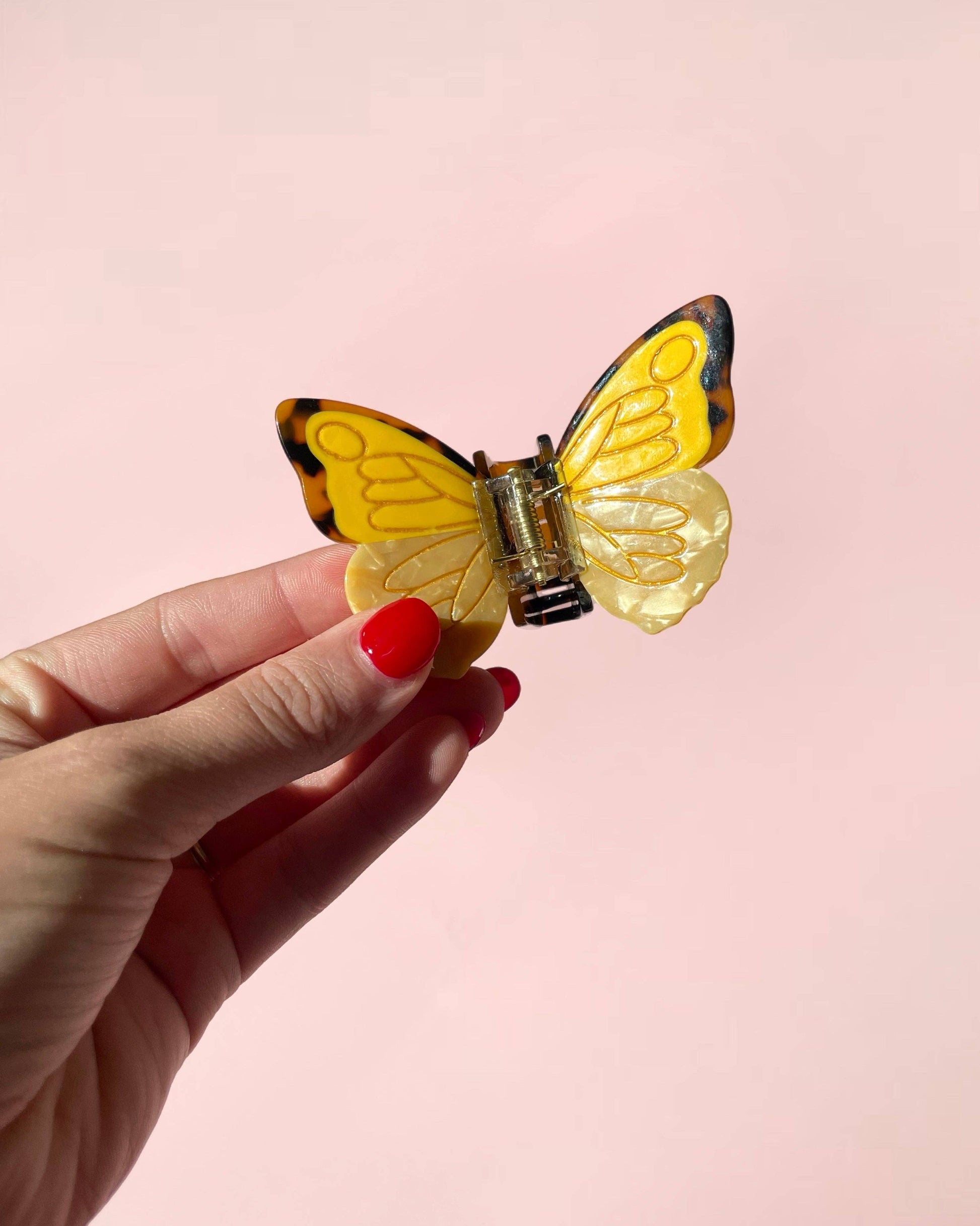 Butterfly-shaped hair clip held by a hand with red nail polish against a pink background