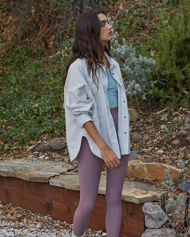 Woman standing outdoors in a natural setting wearing a light shirt, purple leggings, and gray shoes.