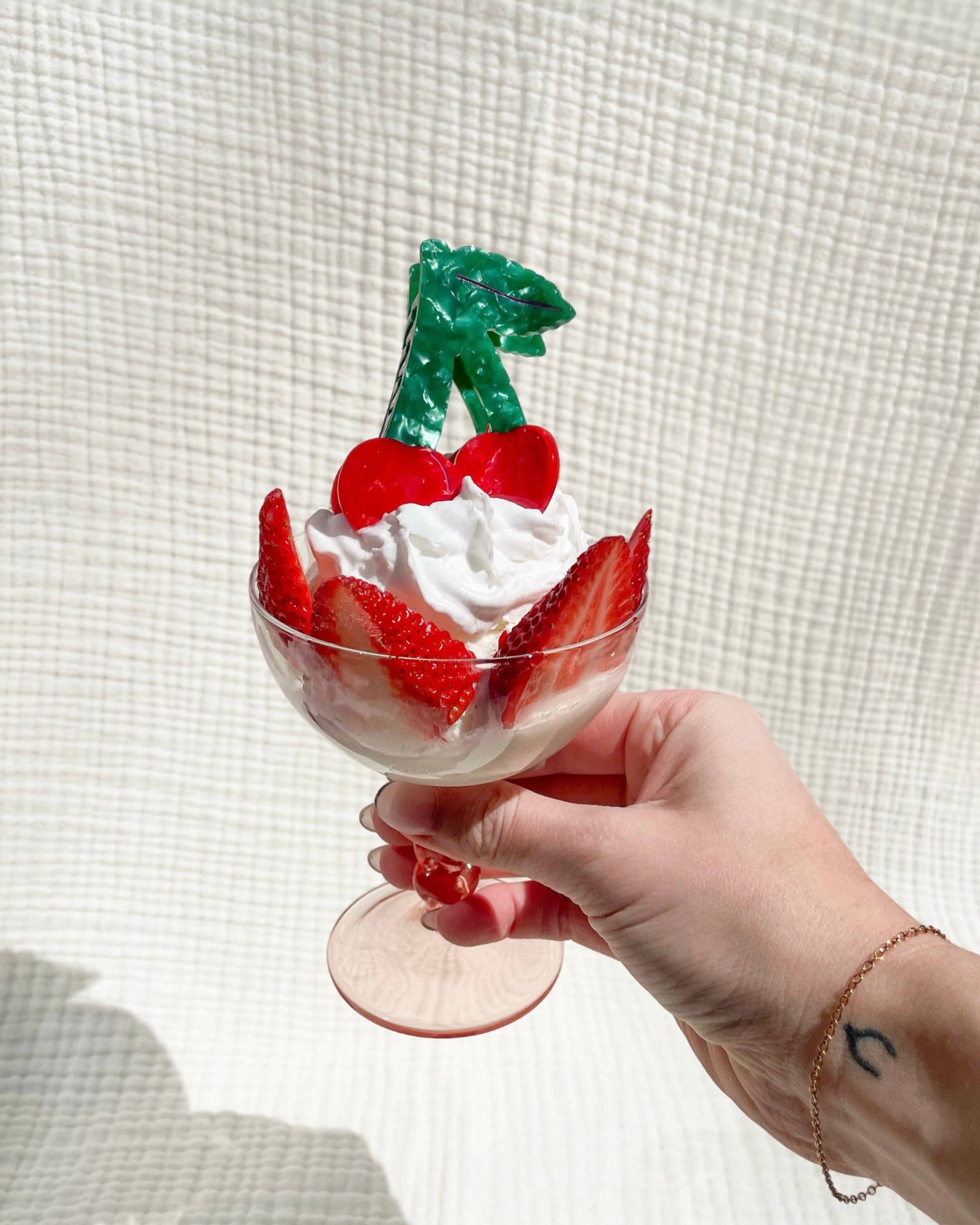 Hand holding a dessert with whipped cream, strawberries, and a cherry-shaped hair clip decoration against a textured white background.