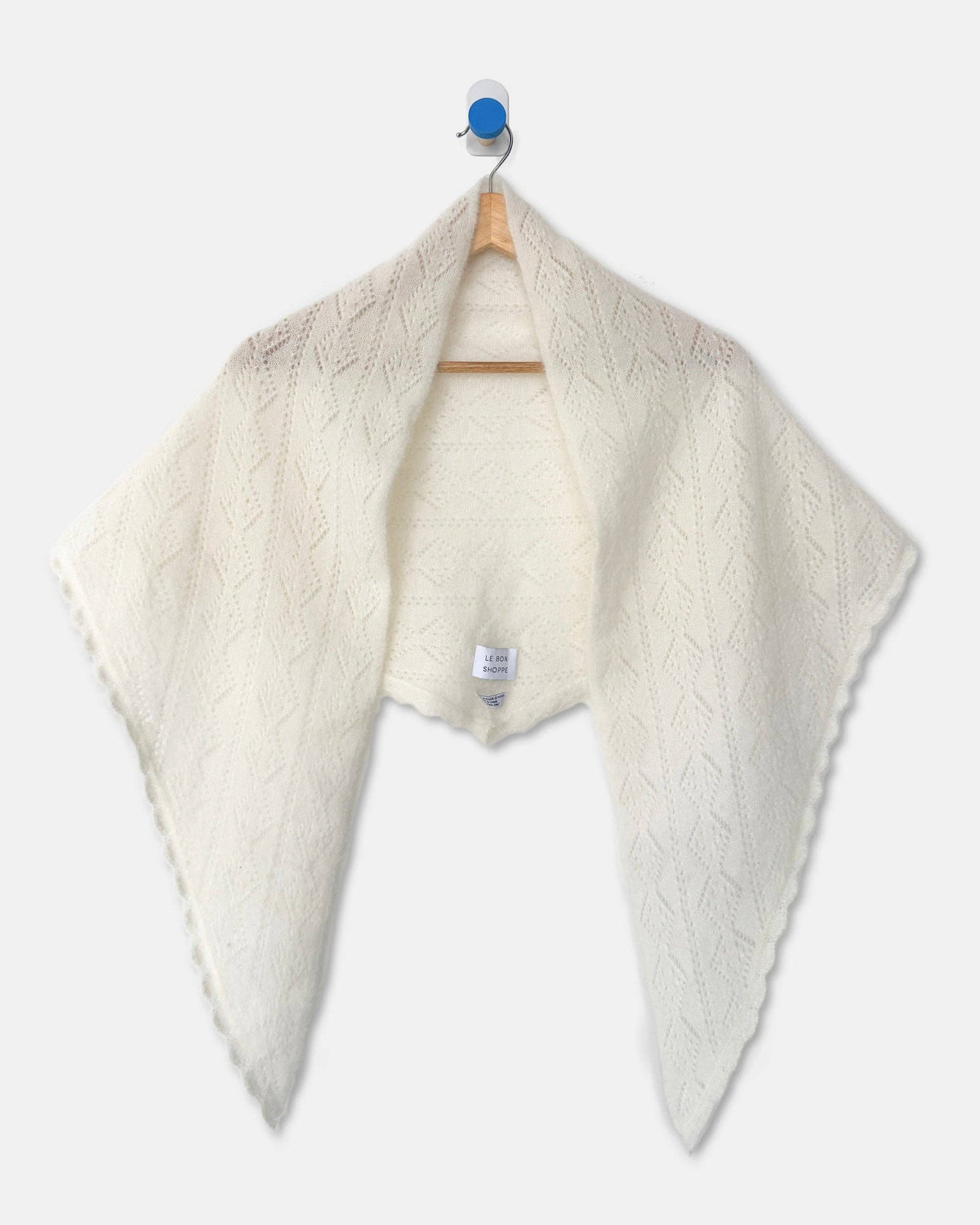 An off-white cable-knit shawl with a scalloped edge, hanging on a wooden hanger against a light gray background.