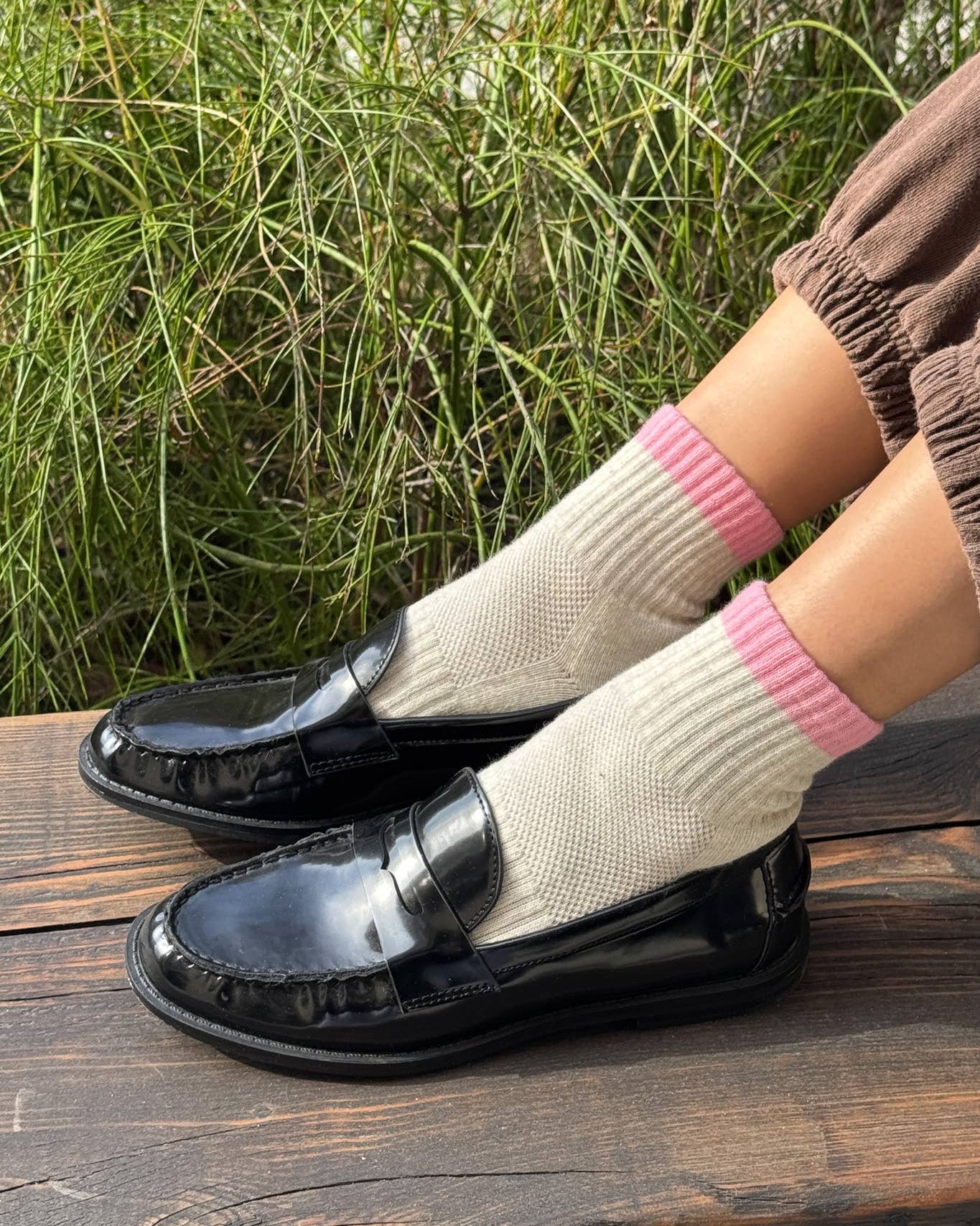 Person wearing black shoes and white socks with pink accents sitting on a wooden surface with greenery in the background.