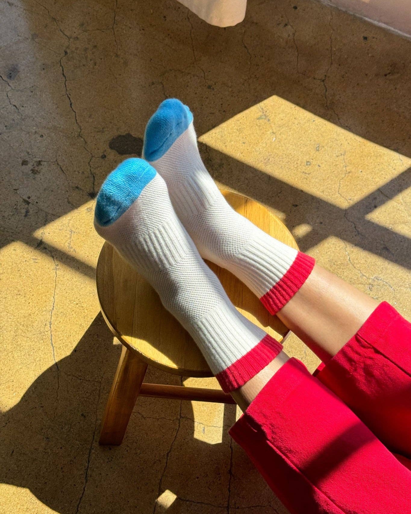White socks with blue toes and red cuffs on a wooden stool with a person wearing red pants.