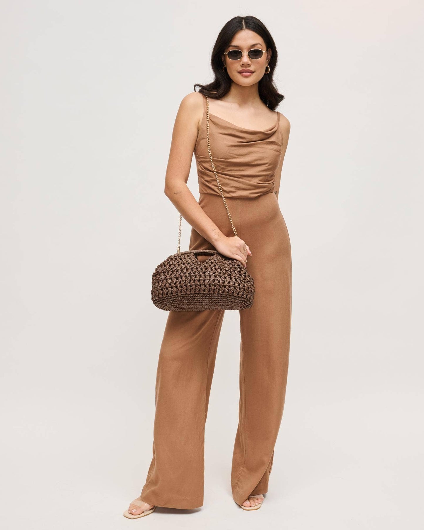 Woman in a brown jumpsuit holding a woven handbag against a plain background