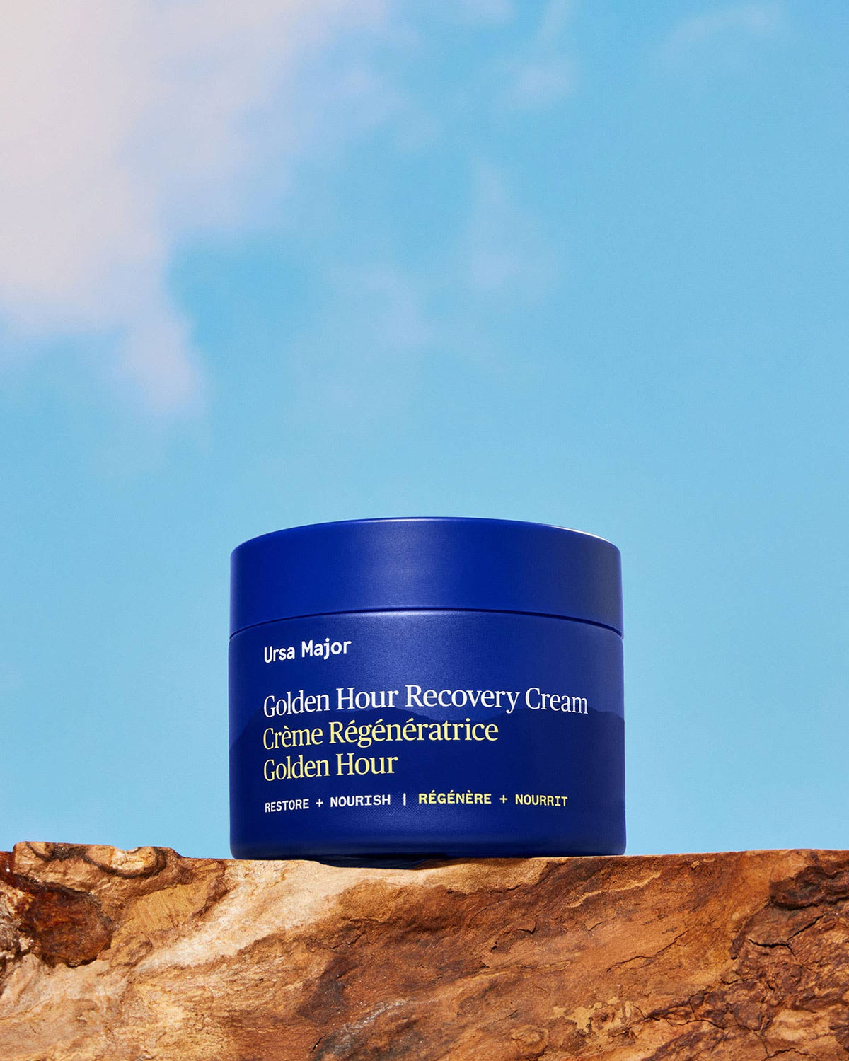 Blue jar of Ursa Major Golden Hour Recovery Cream on a wooden surface with a blue sky background