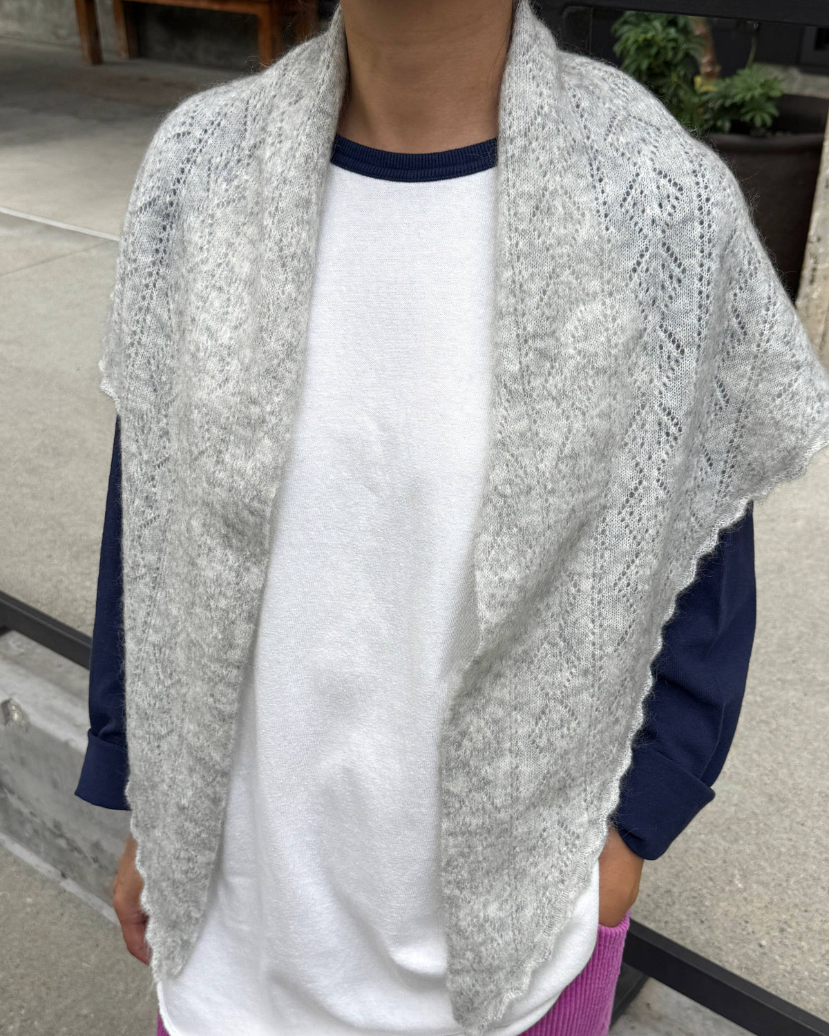 A person wearing a light gray open-front knit shawl or vest over a white ringer T-shirt with navy sleeves, paired with magenta corduroy pants, photographed outdoors on a concrete surface.