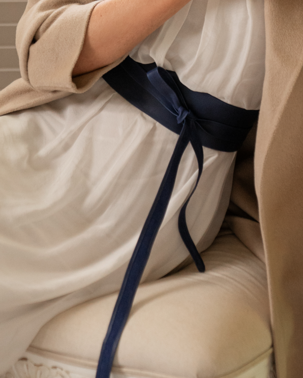 Close-up of a person wearing a beige dress with a navy blue belt sitting on a beige chair.