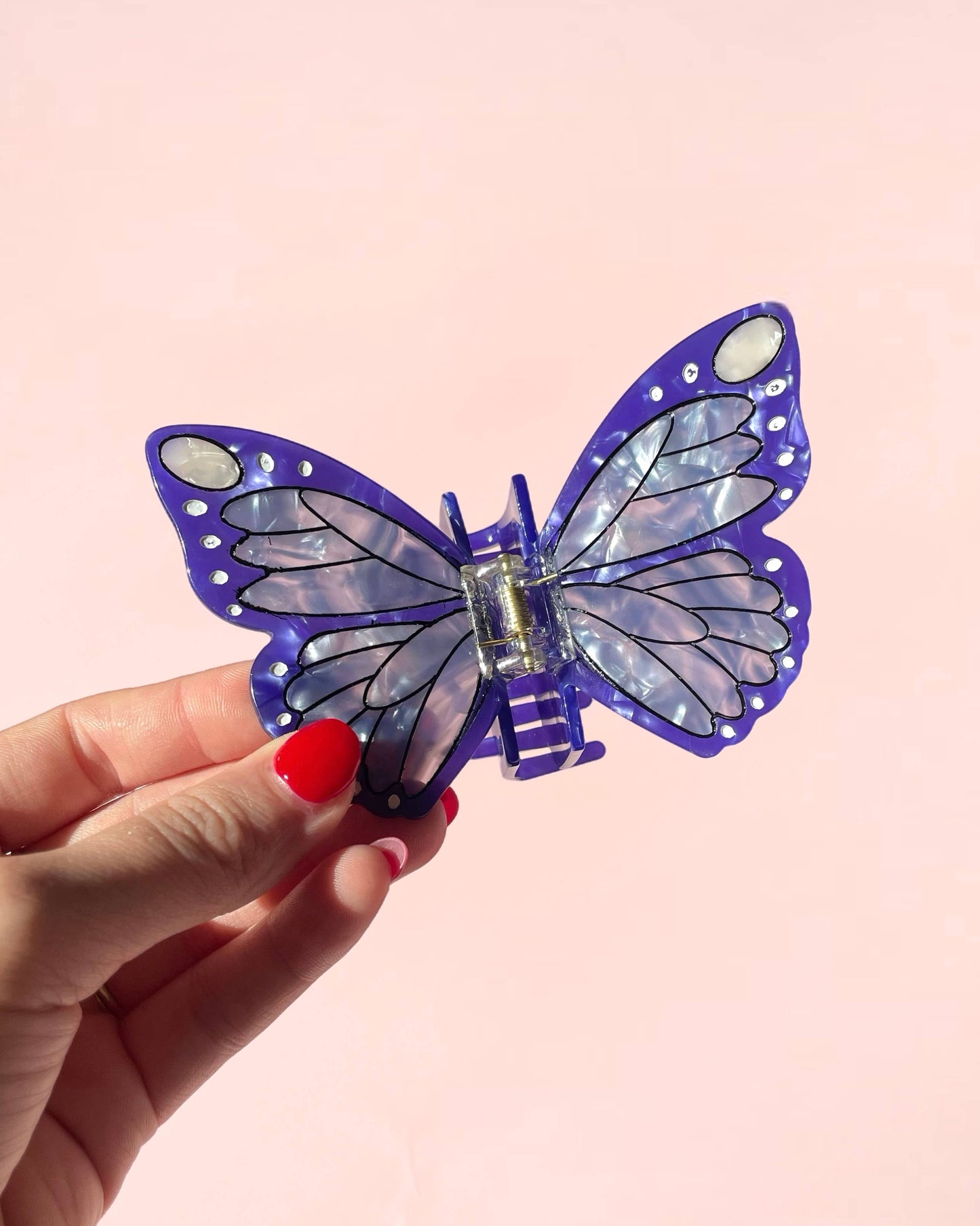 Butterfly-shaped hair clip held by a hand with red nail polish against a pink background