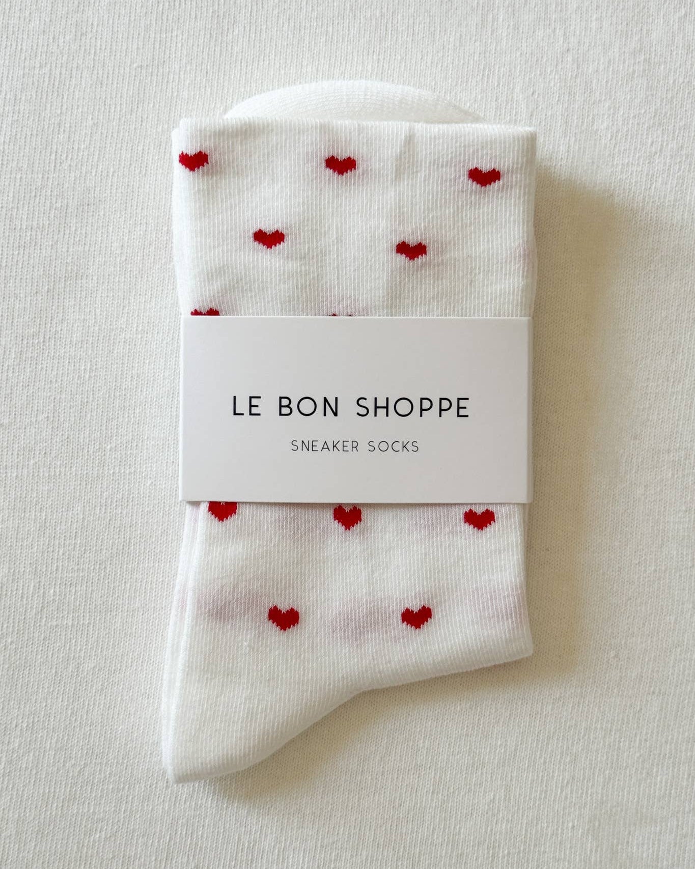 White socks with red heart patterns in packaging labeled 'Le Bon Shoppe' on a light gray background