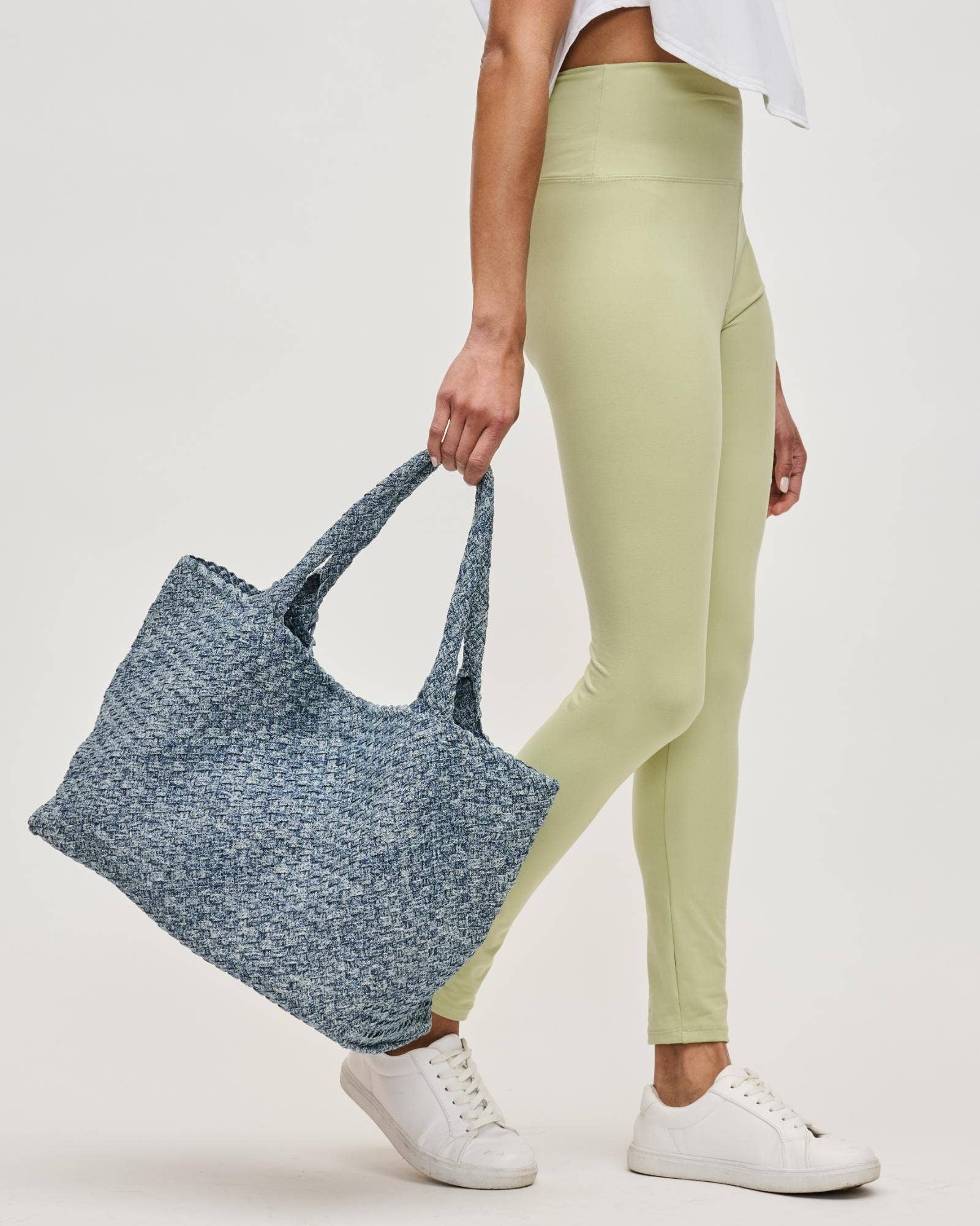 Person holding a blue woven tote bag wearing light green leggings and white sneakers on a plain background