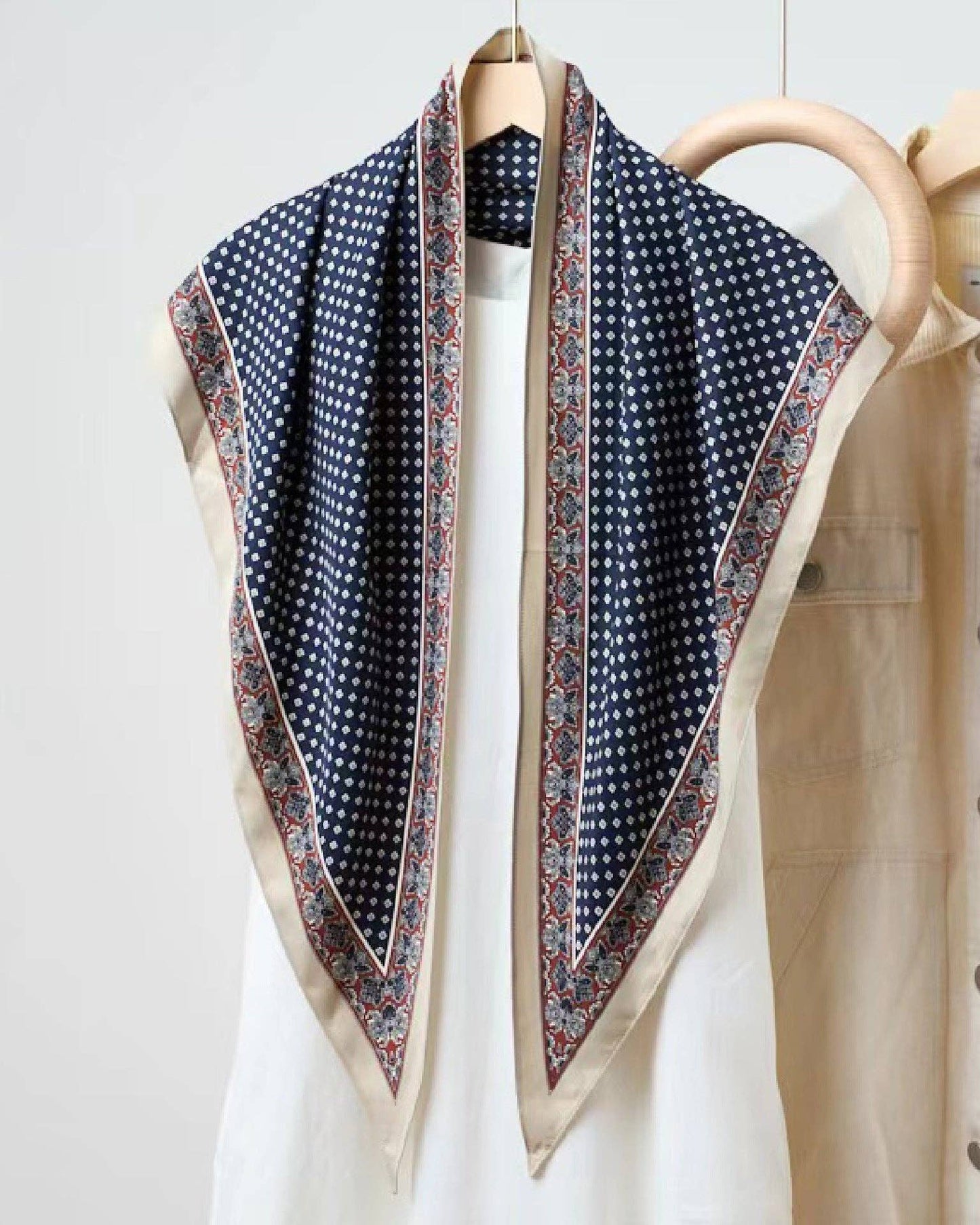 Navy polka dot scarf draped over a white shirt with beige jacket on a wooden hanger against a light background.