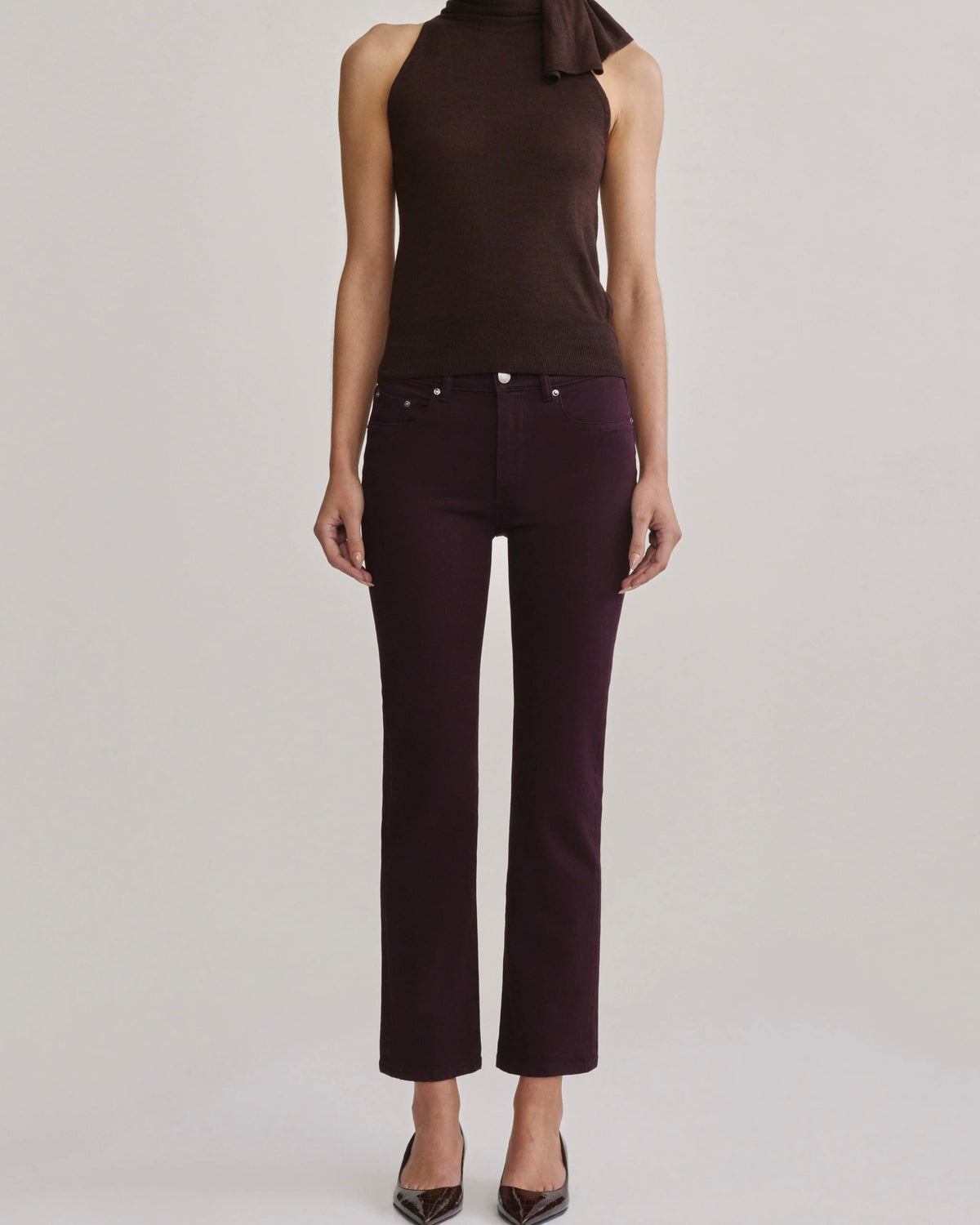 A model is shown from the neck down wearing dark brown sleeveless knit top with a tie-neck detail and dark plum purple cropped pants, paired with brown pointed-toe heels, against a light gray studio background.