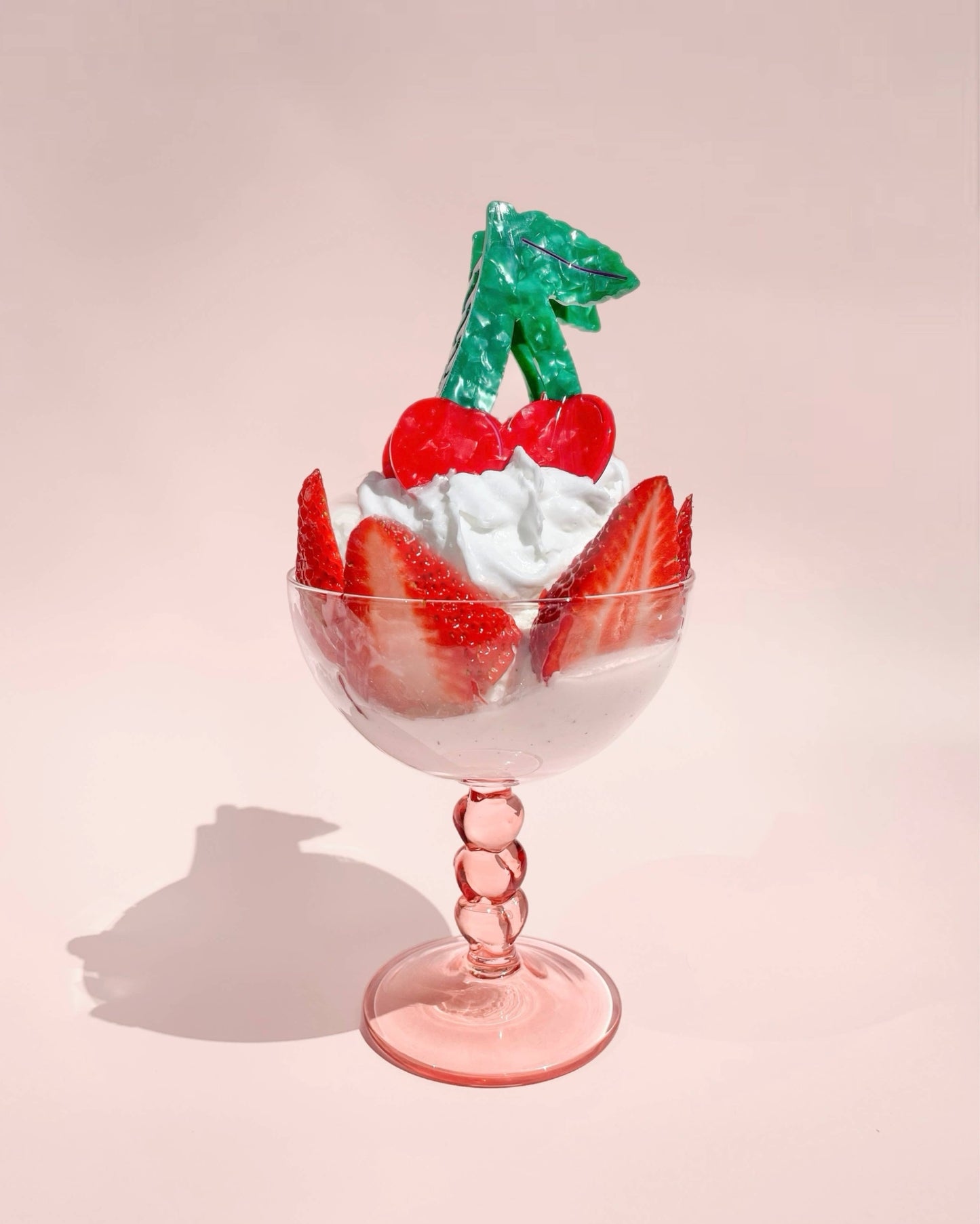 Cherry shaped hair clip laying on a light pink colored glass on a light pink background