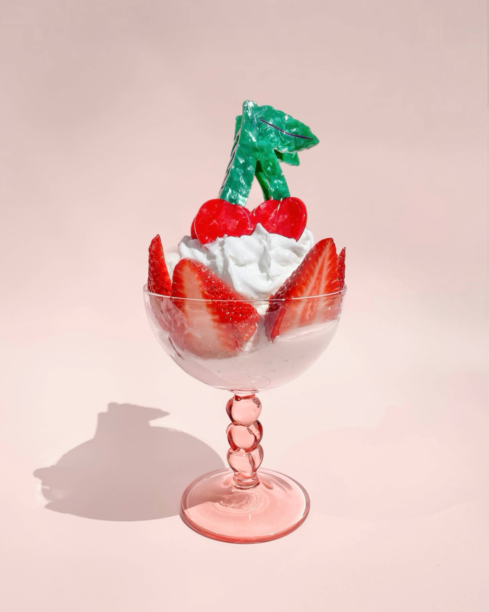 Cherry shaped hair clip laying on a light pink colored glass on a light pink background