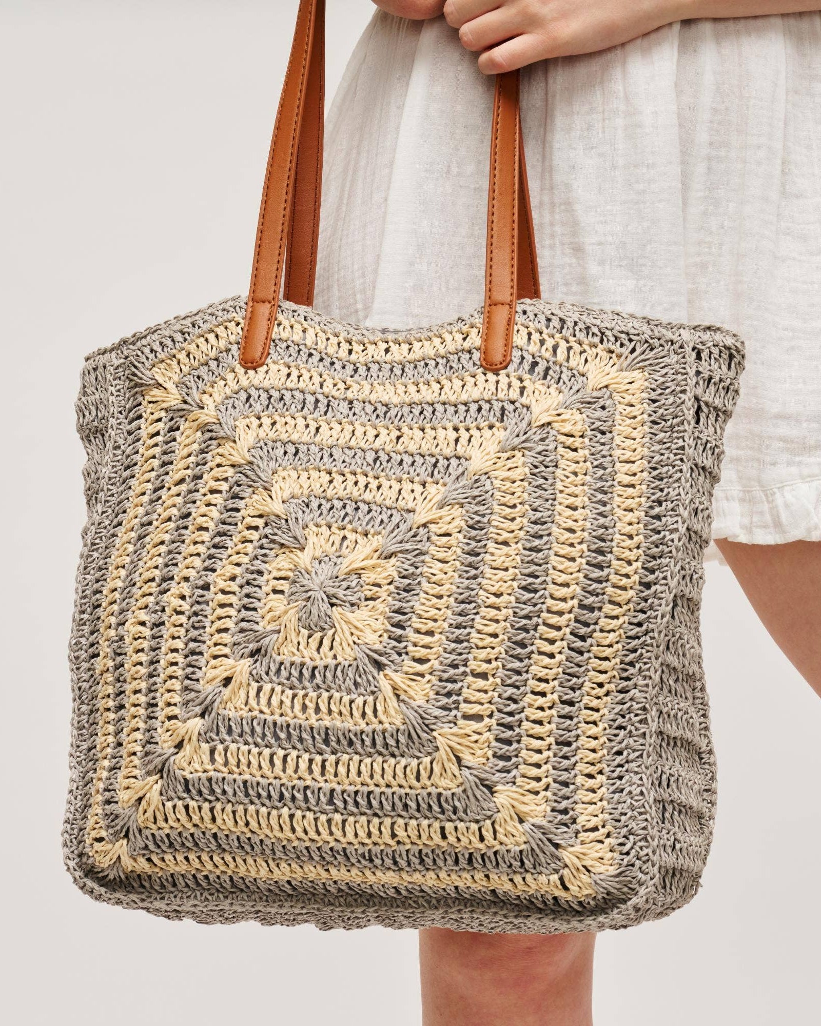 Woven tote bag with brown handles held by a person on a light gray background