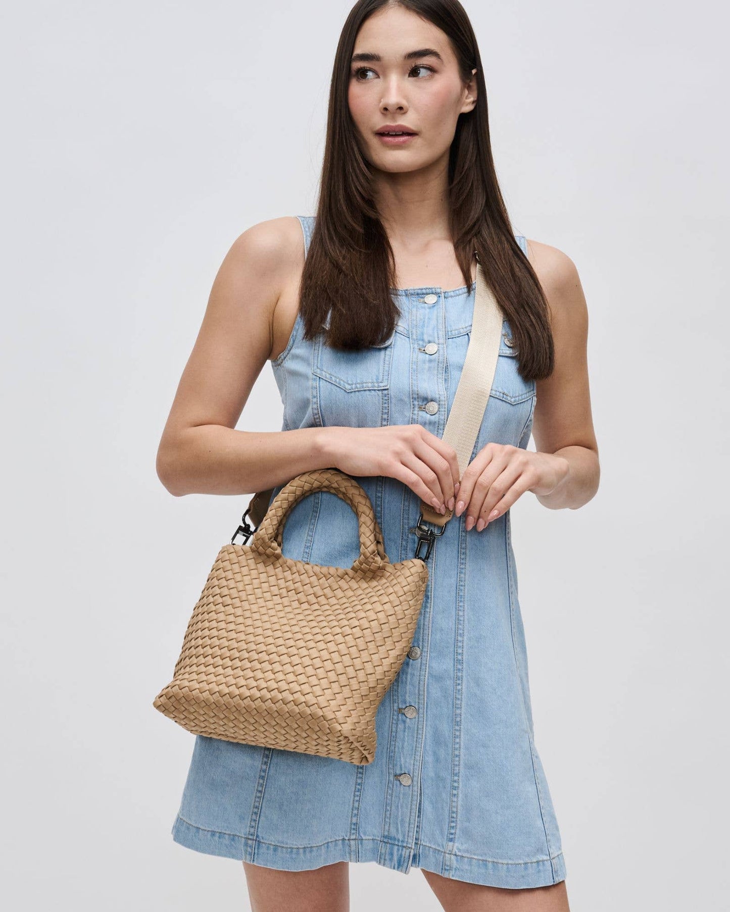 Woman holding a woven handbag wearing a denim dress on a plain background