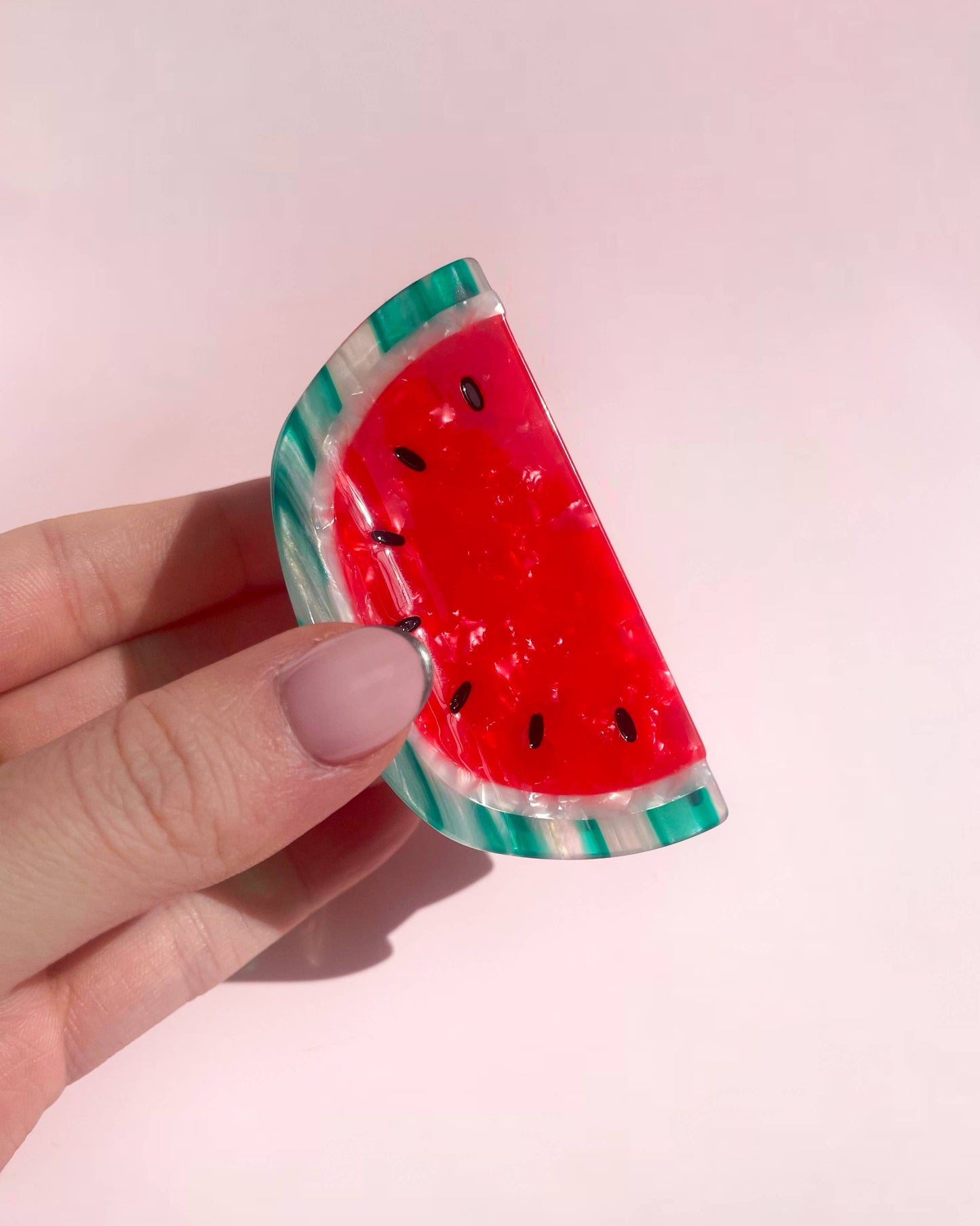 Hand holding a watermelon-shaped hair clip against a pink background