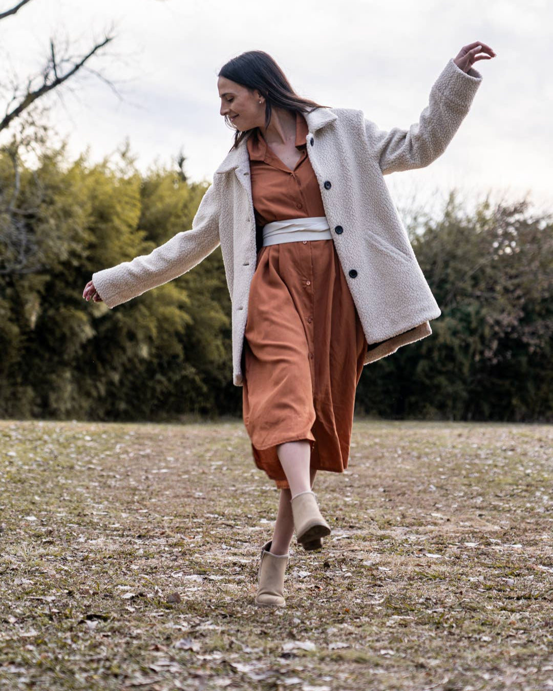 Woman in a white coat and orange dress walking in a field with trees in the background