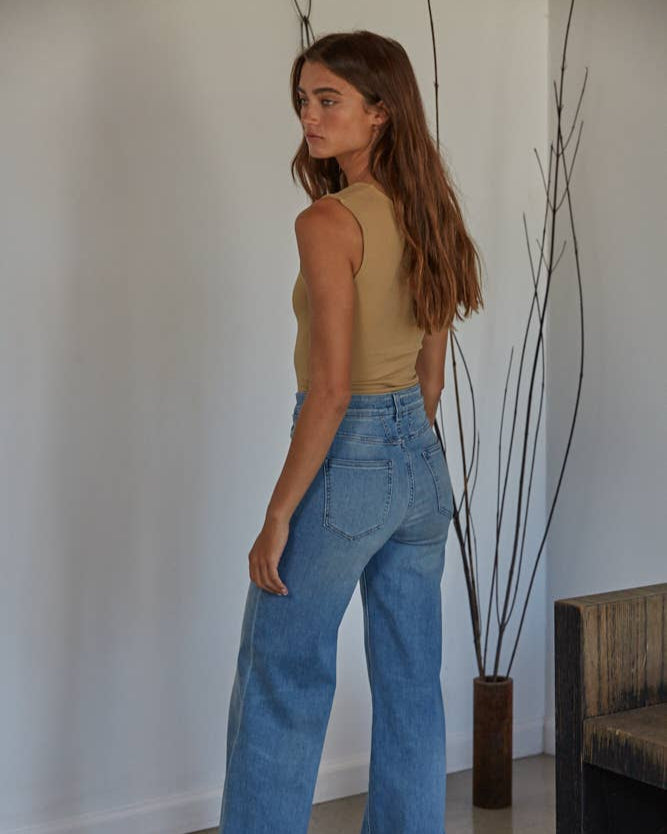 Model wearing a beige tank top with blue jeans in front of a minimalist background.