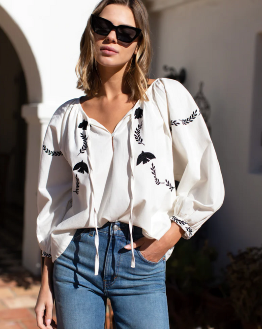 Model wearing Emerson Fry Lucy Dove Embroidery Blouse in Salt Organic wearing jeans and black sunglasses standing outside in front of house on a sunny day