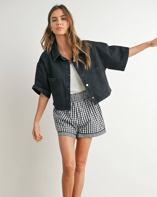 Woman wearing a black denim jacket, white shirt, checkered shorts, and black sneakers against a plain background