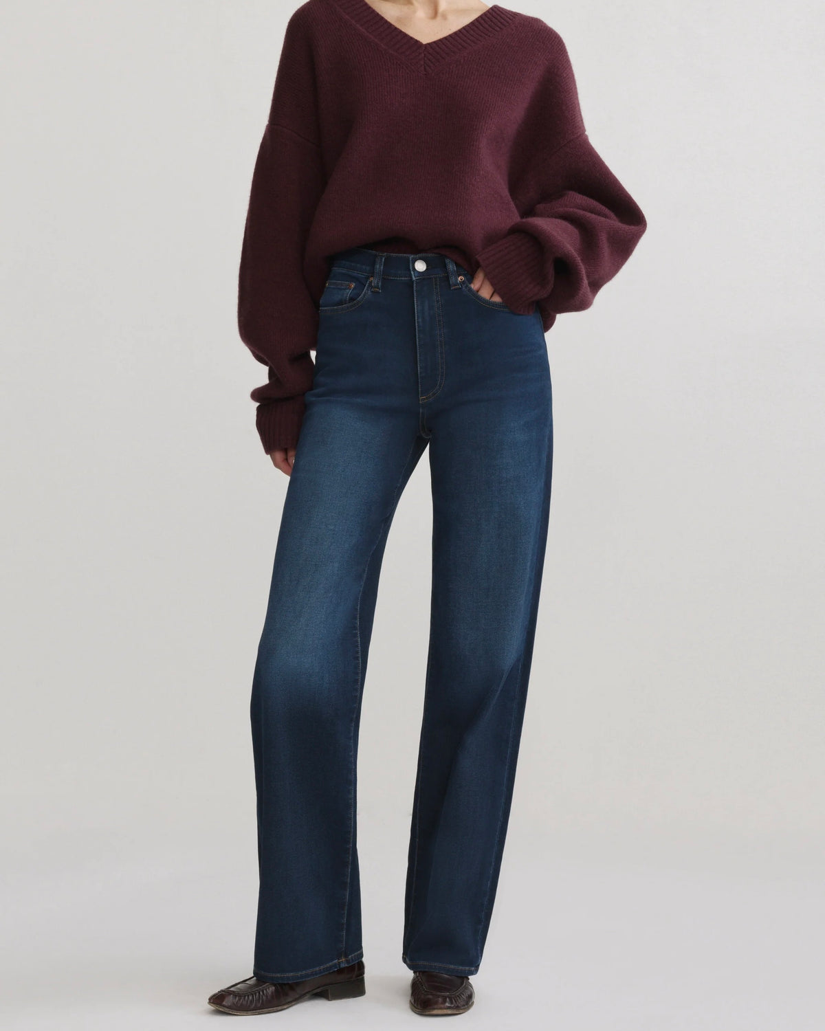 A model wearing dark blue straight-leg jeans and a dark burgundy sweater, standing against a light gray studio background.