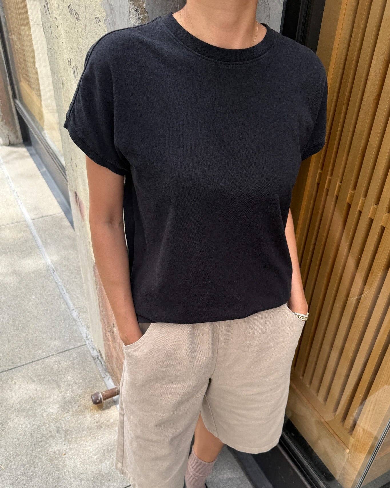 Person wearing a black t-shirt and beige pants standing on a sidewalk.
