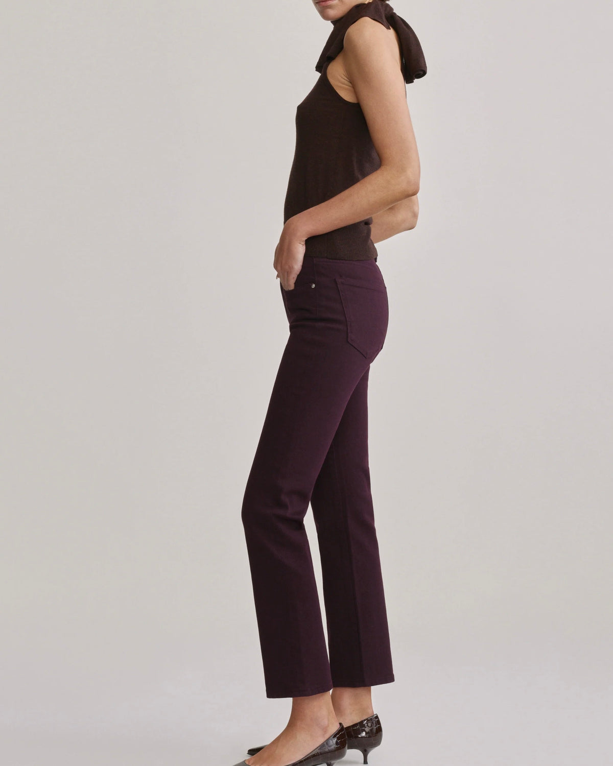 A side-profile view of a model wearing dark plum-colored cropped pants with a dark brown sleeveless top, set against a plain light gray studio background.