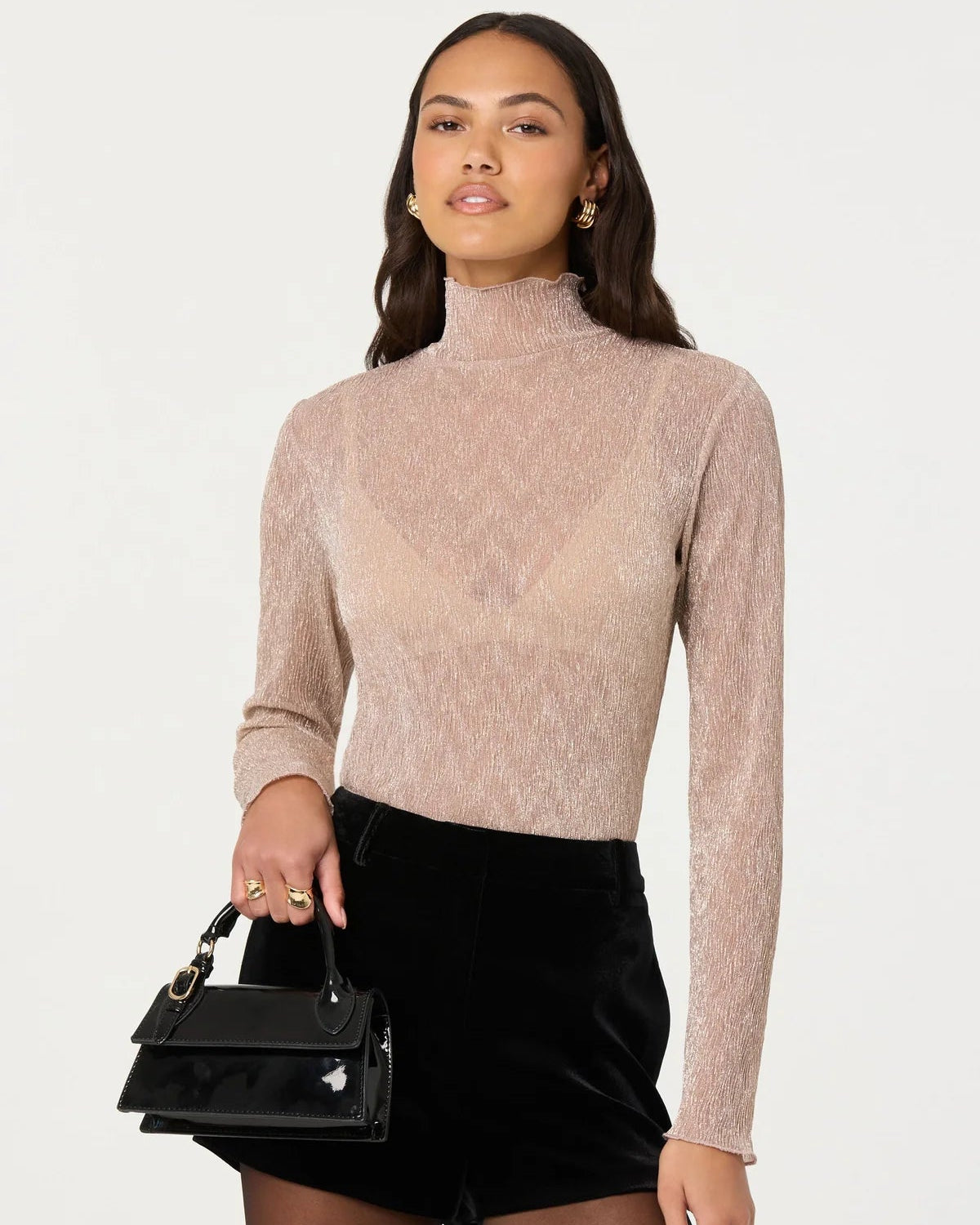 Woman wearing a beige turtleneck sweater and black shorts holding a black handbag on a white background