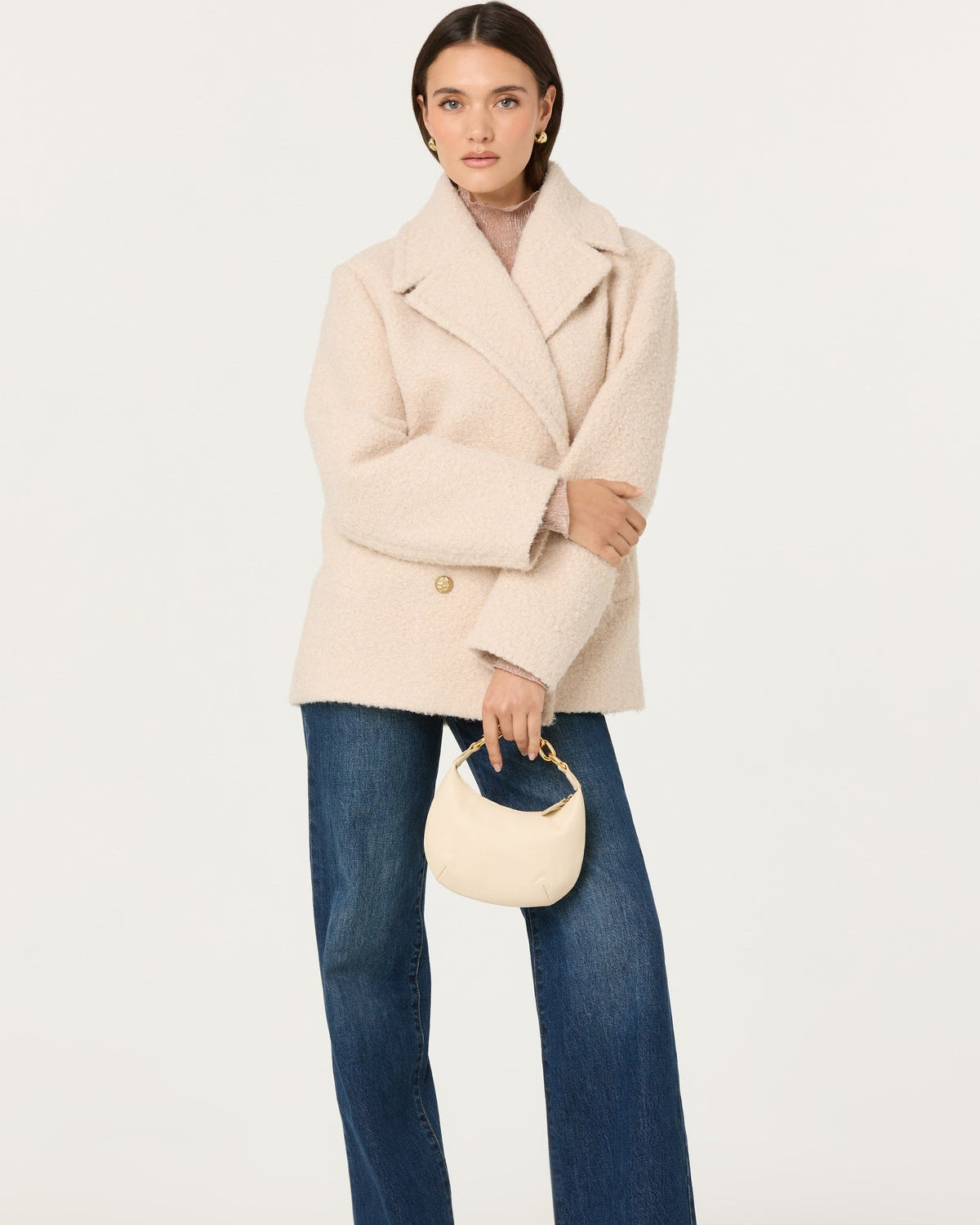 Woman wearing a beige coat and blue jeans, holding a cream handbag on a white background
