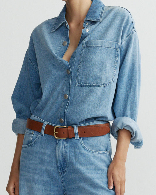 Person wearing a blue denim shirt and jeans with a brown belt on a light gray background