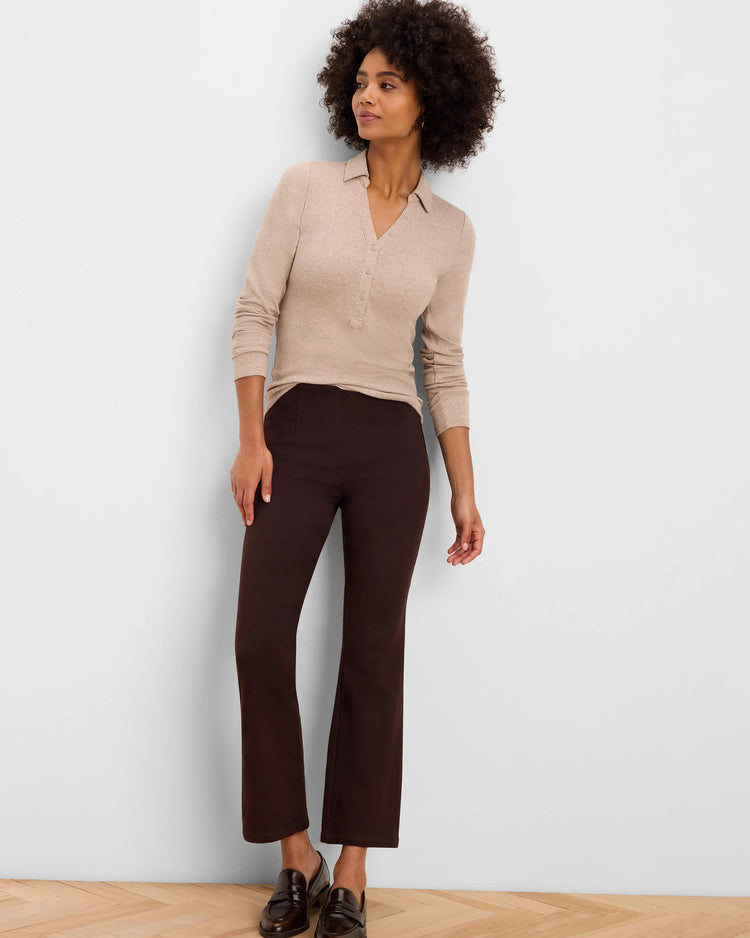 A woman with curly dark hair stands against a plain light gray wall wearing a beige long-sleeve knit polo shirt, dark brown cropped trousers, and dark brown leather loafers.
