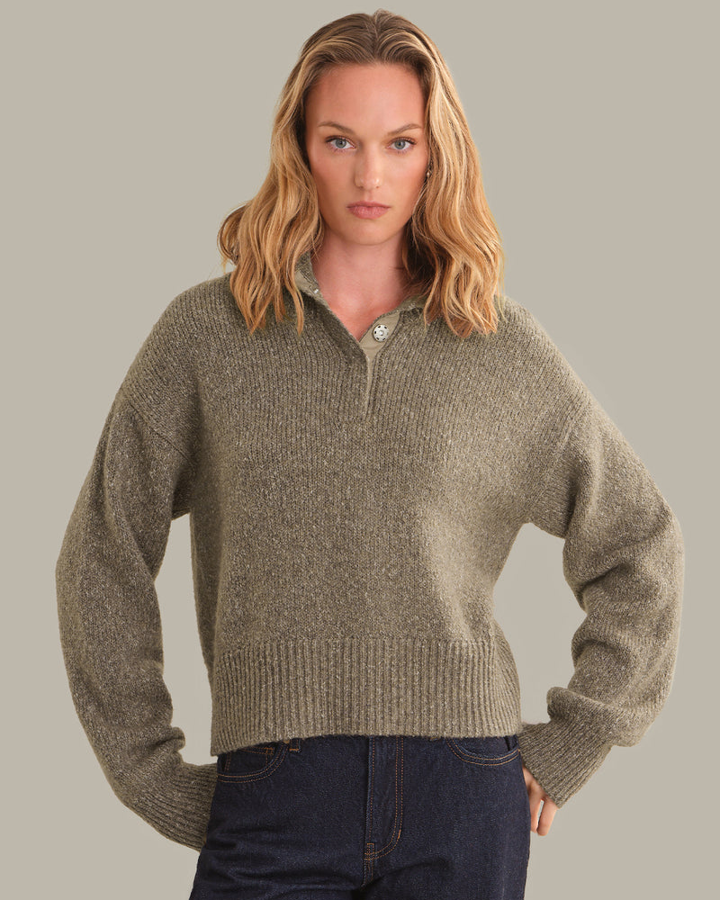 A woman modeling a cropped, ribbed knit half-zip sweater in a heathered taupe-brown color, paired with dark blue jeans, against a solid light gray background.